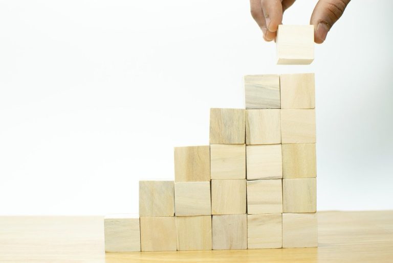 Continuous improvement A hand places a wooden block on a step-like arrangement of stacked blocks.